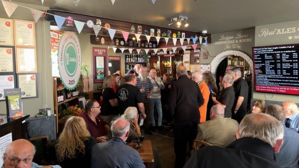 Customers standing inside the bar area of the Blackfriars Tavern, having raised a toast, gathered together in conversation. Bunting is hanging from the ceiling, with awards, menus and other signage on the walls.