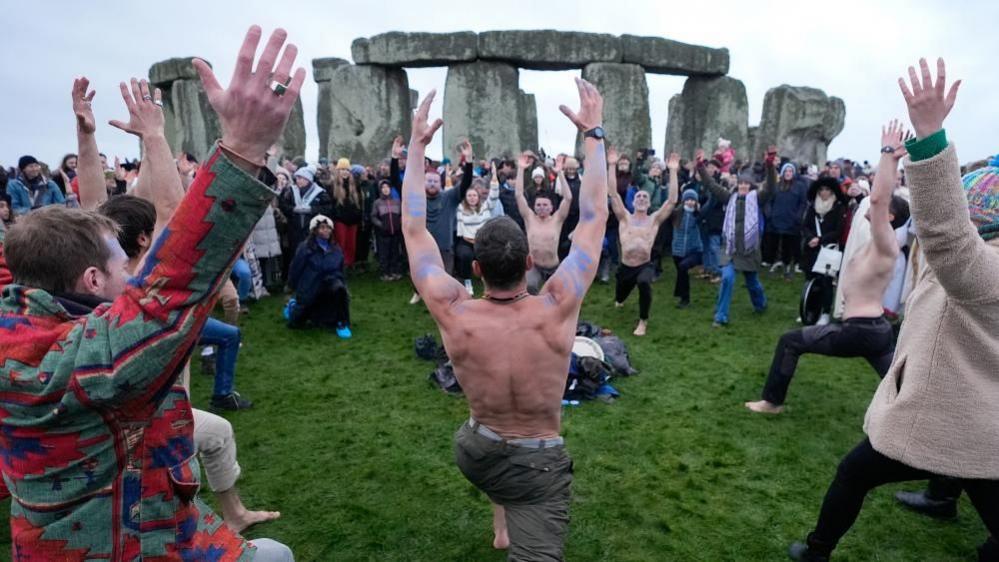 A group of people in a lunging pose with their hands raised above their heads, in front of the Stonehenge stones