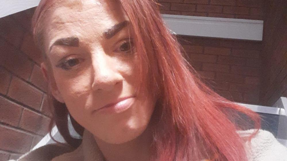 A selfie of a woman who has red hair. She is sitting outside in the sunlight in front of a brick wall. She is smiling.