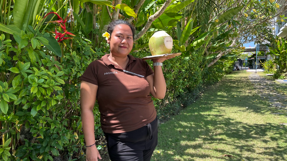 Ni Kadek Sintya in a brown polo shirt, holding a coconut on a plate. There is a yellow frangipani in her hair and she is smiling.