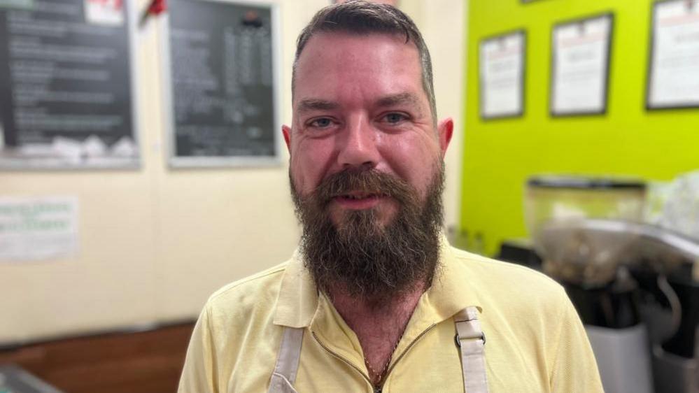 Nigel Lyons smiling at the camera and wearing a pastel lemon top with cream coloured apron and with his coffee shop blurred in background