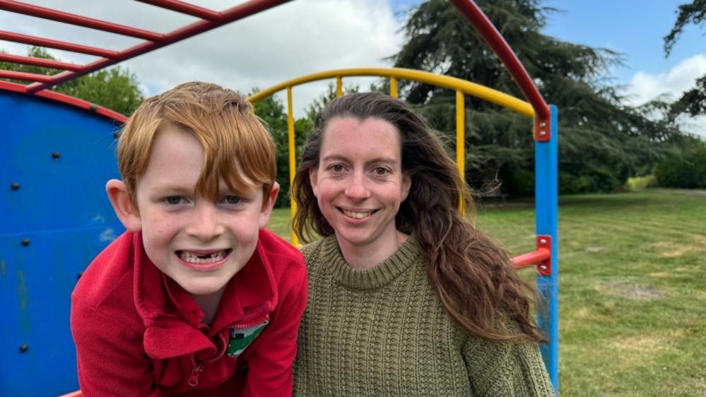 Jess Royal and her son on the play equipment at Cantley Village Hall. She has long brown hair, and is smiling, wearing a green knitted jersey. Her son has ginger hair, has a gap-toothed smile as his adult teeth grow and he is wearing a red fleece embroidered with the school logo. The play equipment is made of red and yellow iron bars, with a blue board to the left of the image, with grass and trees to the right and in the backdrop.