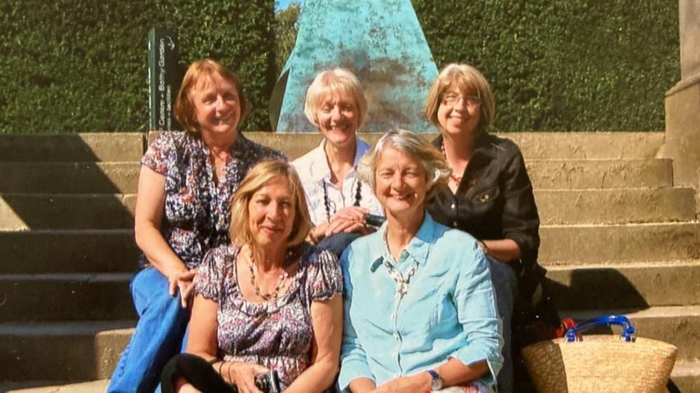 Five women sit on steps. Three women on the back row and two in the front. They are wearing jeans and blouses and one has glasses. 