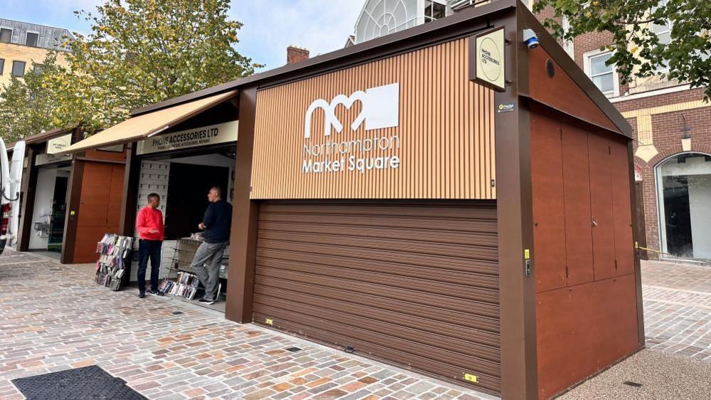 Wood-coloured market stall with brown shutter and the Northampton market logo. Two people stand at an adjacent stall. There are new brown and grey paving stones on the ground.