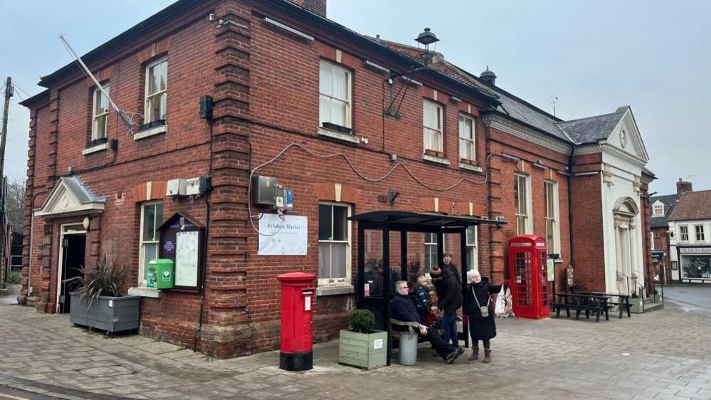 Aylsham Town Hall, a red-brick Georgian building with decorated features and a grand entrance to the right of the image. Outside it are a red pillar box, a bus shelter and a red K6 British telephone box.