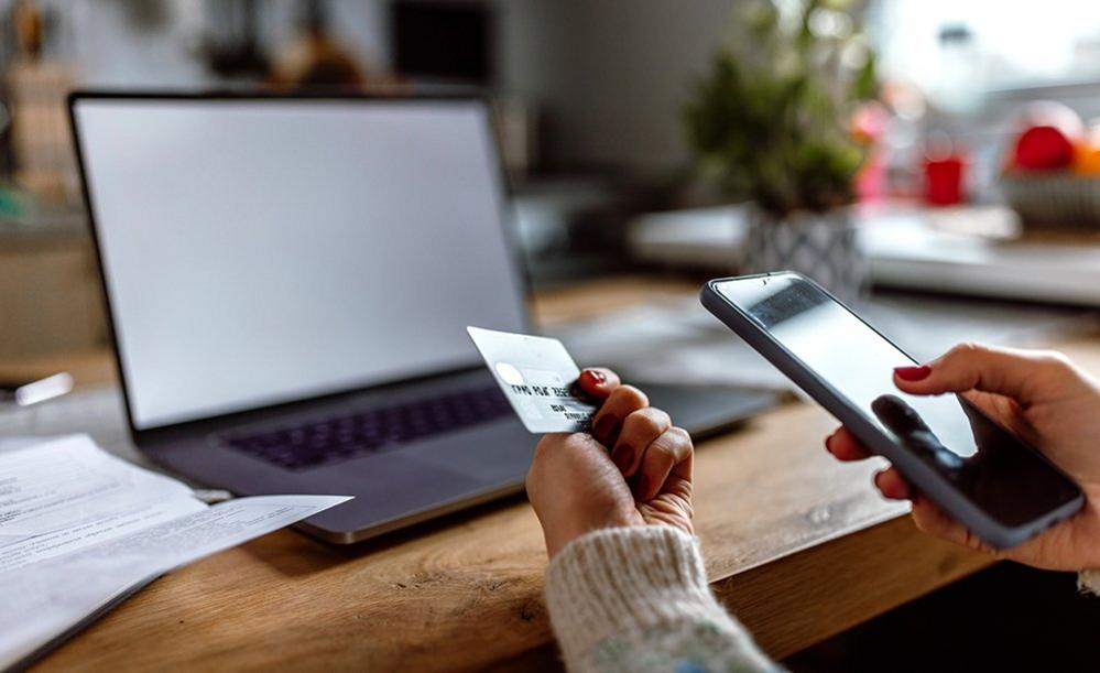 Hands holding smartphone and credit card in front of laptop on kitchen table at home