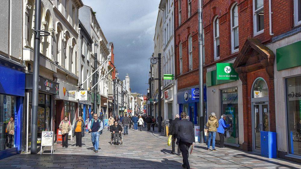 A view of Strand Street with shoppers walking through the centre of Douglas high street on a sunny day. 