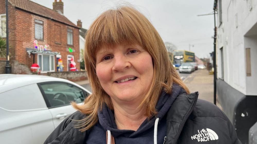 Julie Smith has auburn hair, is wearing a blue hoodie and a black puffa style jacket. She is standing next to a filthy white van parked on Acle High Street.