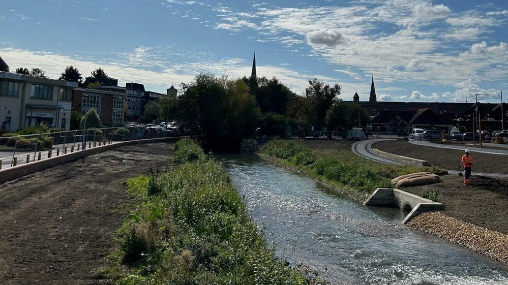 A river with large expanded banks to allow flooding with properties to the left and a road to the right