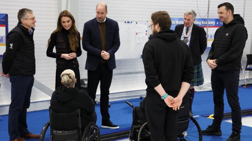 Princi dhe princesha qëndrojnë në një turmë njerëzish në pistën e kaçurlingut duke biseduar.