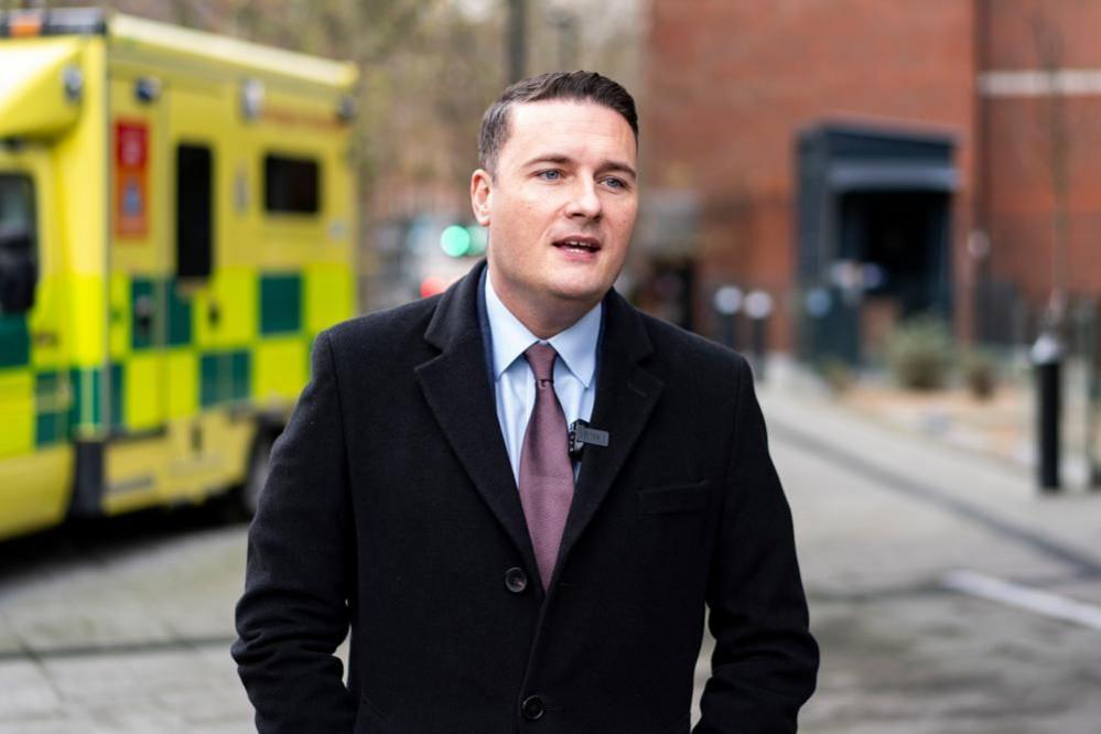 Health Secretary Wes Streeting is seen speaking to reporters outside a hospital. He is wearing a black coat and purple tie. Over his right shoulder an ambulance is seen parked.