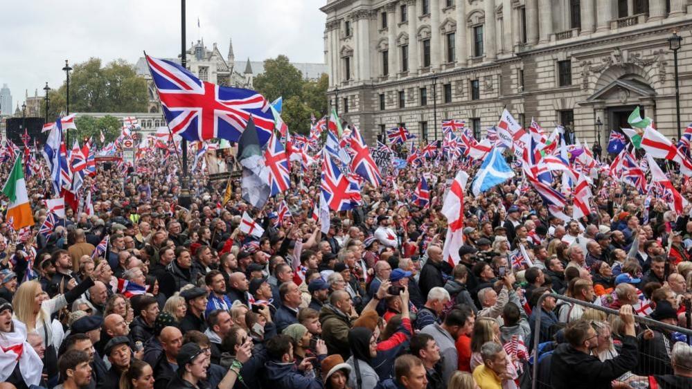 Large crowds fill Whitehall in central London with many seen waving union jacks and St George's flags, with grand Whitehall cream-coloured buildings seen in the background.