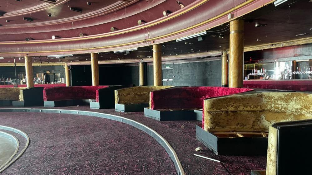 The interior of the Ocean Room, showing scalloped-shaped banquette seating, upholstered alternately in red and gold velour. The ceiling has contours in the Art Deco style, and the floor is raked down towards the dance floor. The bars are visible on the back wall of the building.