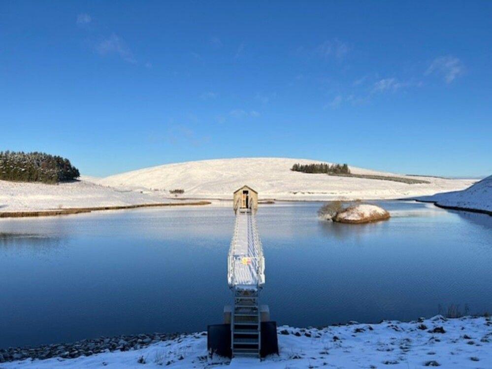 North Esk Reservoir in the Pentland Hills. A blue reservoir surrounded by low snowy hills and a blue sky. A bridge stretches out to a little house at the other end of the reservoir.