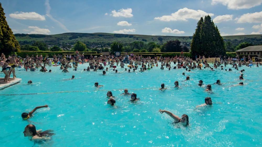 Children swim in an outdoor pool on a sunny day. There are moors visible in the background