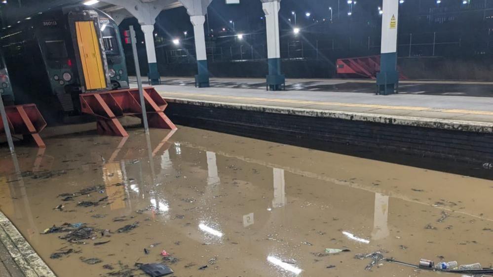 A flooded station, with water and debris on the platform. There is a train at the platform