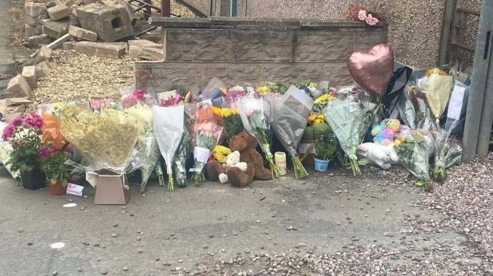 Around 30 bouquets of flowers lie beside a broken stone wall. The floral tribute also includes two teddy bears, a heart shaped balloon and a candle. Stone rubble is visible in the background.