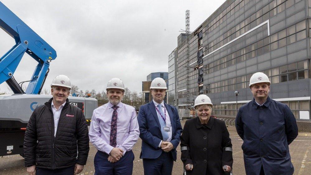 Four men and one woman are standing in a line in front of a large office block. It is grey and covered in glass windows. They are wearing white hard hats and there is a grey and blue construction vehicle behind them. The woman has crutches.