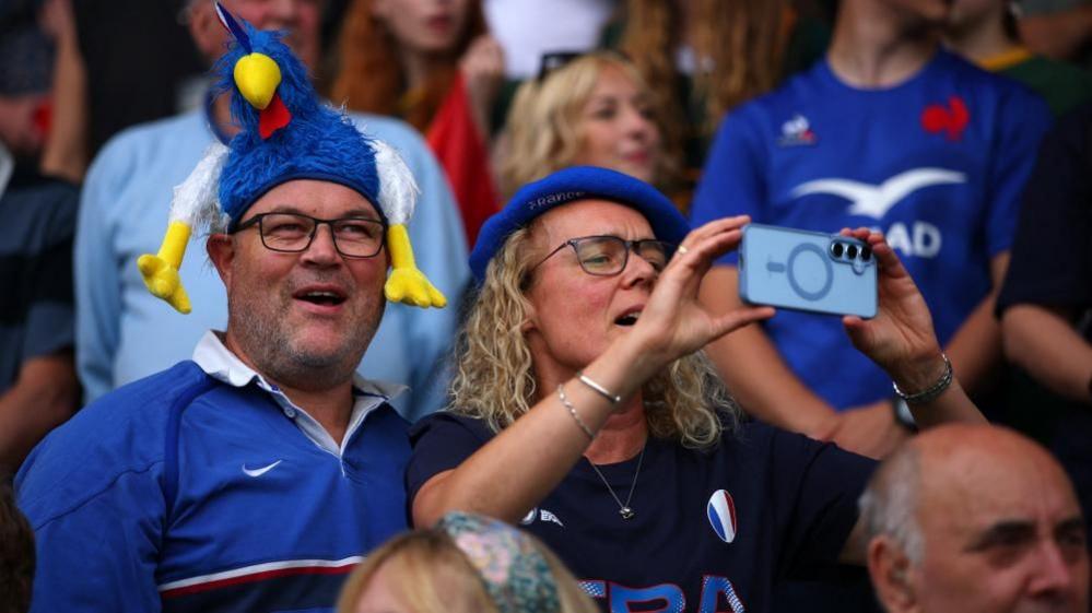Two French fans in the crowd. The man is wearing a blue shirt and a blue cockerel hat, and the woman is wearing a beret.