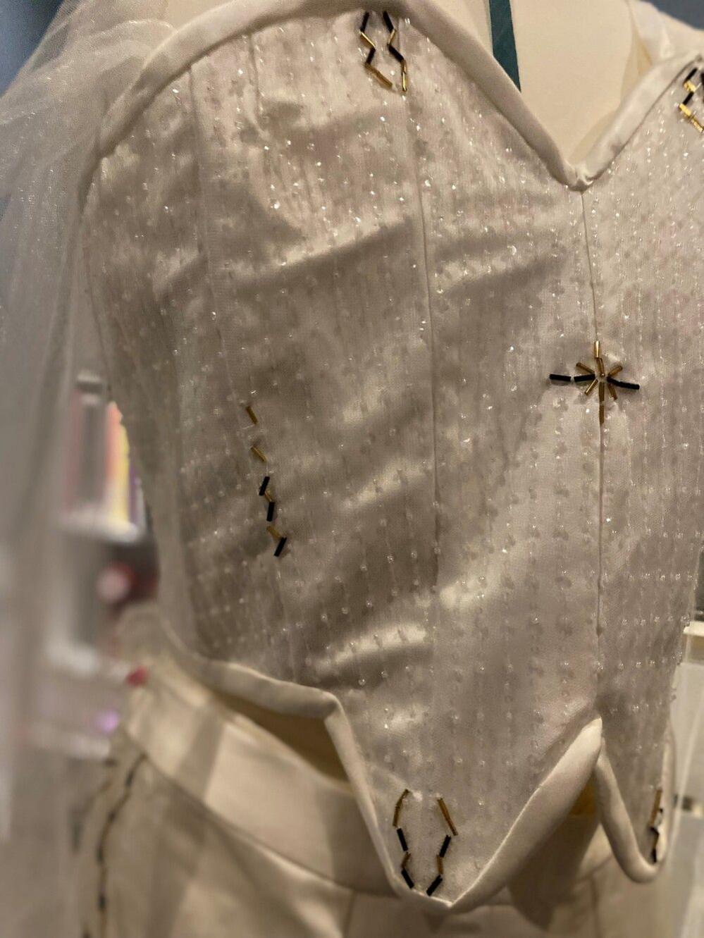 A beaded white corset with a cape in the background and trousers on a mannequin