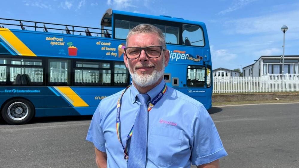 Keith Wakefield has greying hair, and a white beard and moustache, and wears glasses. He is wearing a blue shirt and blue tie, and a rainbow Pride lanyard He is standing next to the Coastal Clipper Cabriolet bus, which is painted in multiple shades of blue, with a yellow diagonal stripe ahead of the rear wheel, sloping towards the back upper deck.