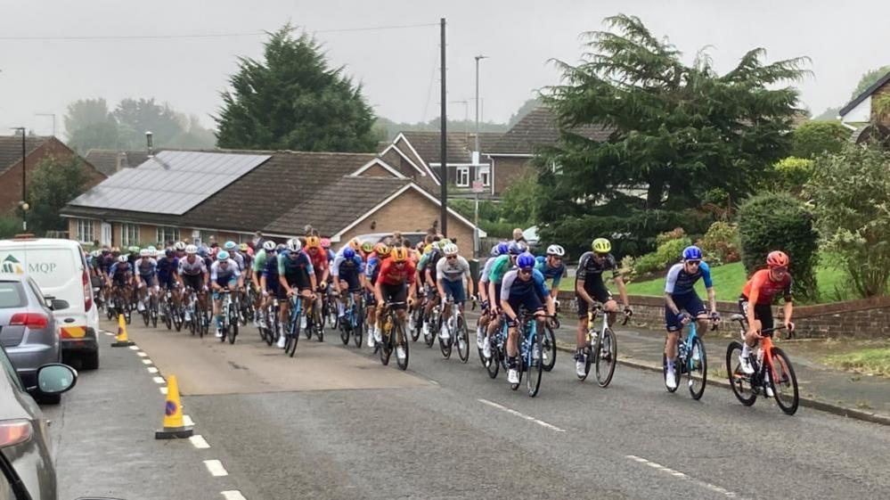 Dozens of cyclists make their way up a road. Police cones and parked cars are visible.