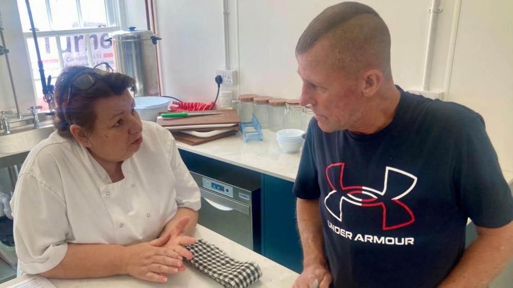 Jon being taught how to cook, looking at a woman in a white chef's coat. Jon has a mohawk and a navy "Under Armour" branded T-shirt. The woman he is talking to has short brown hair and glasses resting on top of her head.