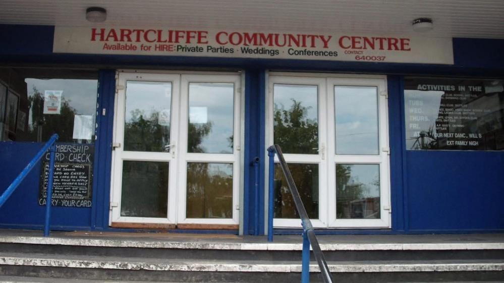 The entrance to Hartcliffe Community Centre. A white sign with red lettering hangs over two white, glazed double doors at the top of some steps. The walls to the centre are blue with other chalk board signage on them.
