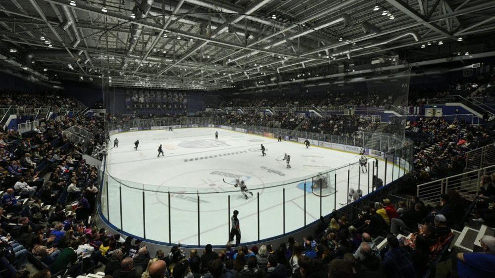 A view from the sands looking onto an ice hockey rink during a match. Two teams are out on the ice skating around, watched by a busy crowd. 