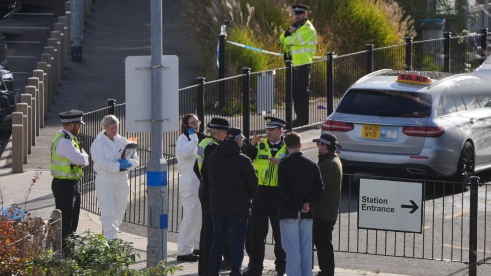 Police officers standing outside railway station entrance. There is crime scene tape in the background 