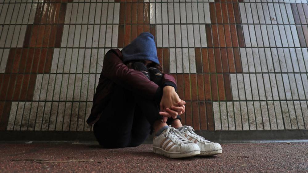 Library shot of child wearing blue hood sitting next to a wall