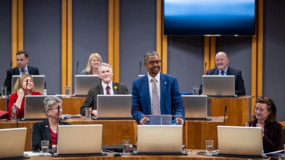 Vaughan Gething yn sefyll ac yn siarad yn y Senedd gyda gwleidyddion eraill yn eistedd o'i cwmpas.