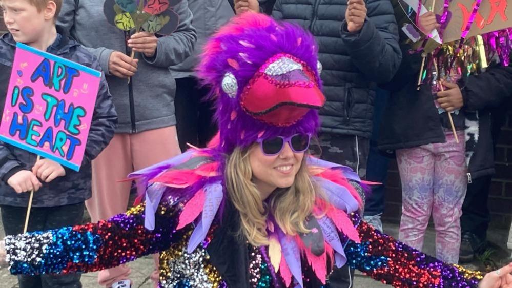 A woman with long blonde hair, wearing purple sun glasses wearing a big purple  fabric crows head and a sparkly outfit made with pink and purple feathers.