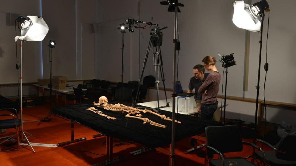 A man and a woman observe a skeleton laid on a black table, with floodlights around the outside