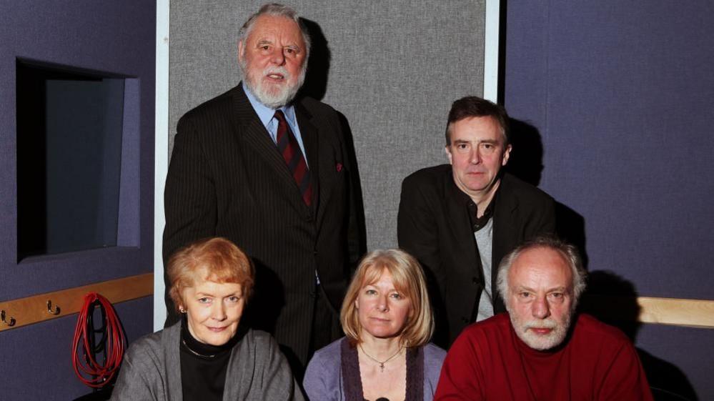 Sir Terry Waite with short grey hair and beard, standing in a radio studio wearing a dark jacket and tie. John McCarthy with short dark hair and wearing a dark jacket is standing to his left. Brian Keenan with short white hair and beard, wearing a red sweater, is setting in front of Mr McCarthy. There are two women sitting next to Mr Keenan - the presenter Sue McGregor and Mr McCarthy's partner, Jill Morrell
