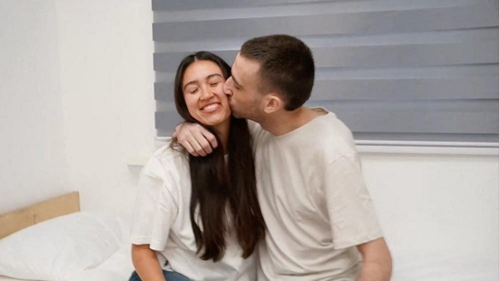 Avinatan Or with short cropped hair wraps his arm around his girlfriend's neck to bring her close and kiss her on the cheek. Both are wearing white T-shirts, and Noa Argamani has long, dark hair and is smiling with her eyes closed