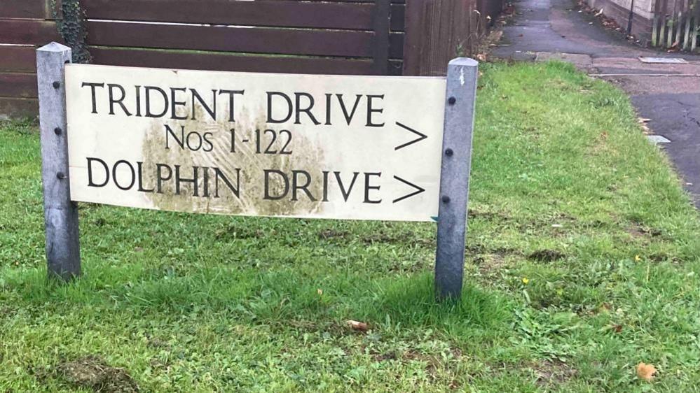 A rectangular white held up by two grey posts sitting in green grass. The words TRIDENT DRIVE Nos 1- 122 and DOLPHIN DRIVE are written in black on the sign.