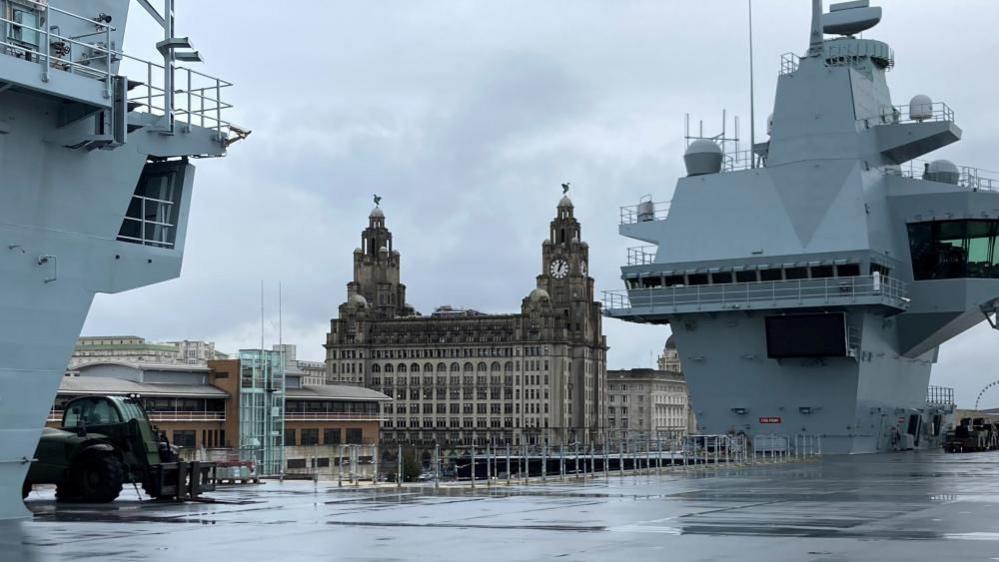 The top deck of a large military ship with a historical building in the background. 