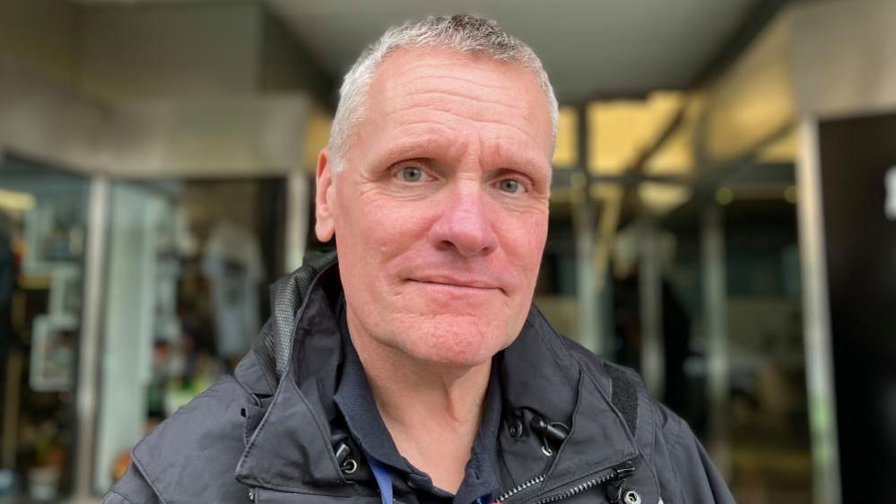 Man smiling outside shop. He is wearing a black coat.