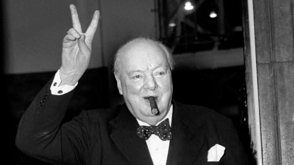 Winston Churchill, in a suit and bow tie and with a cigar in his mouth, makes V for Victory sign with his right hand 