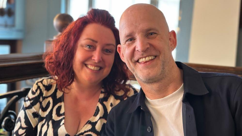 Nicola and Phil Mansford, sitting down, smiling at the camera. Nicole has long red hair, wearing a black and white dress, Phil has a bald head, grey goatee, an earing in one ear and is wearing a white T-shirt and black shirt.