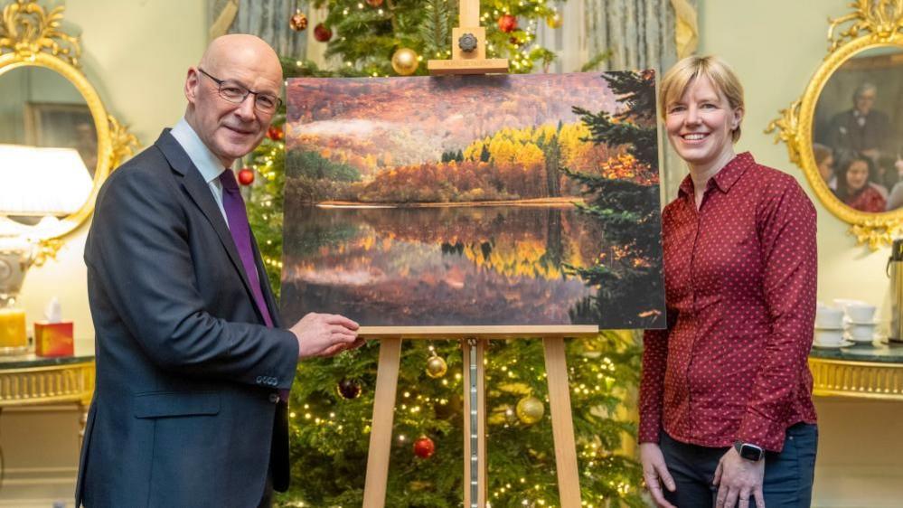 John Swinney standing in front of a Christmas tree with Jane Barlow and the winning photograph - an autumn scene of a loch with trees on a hillside.