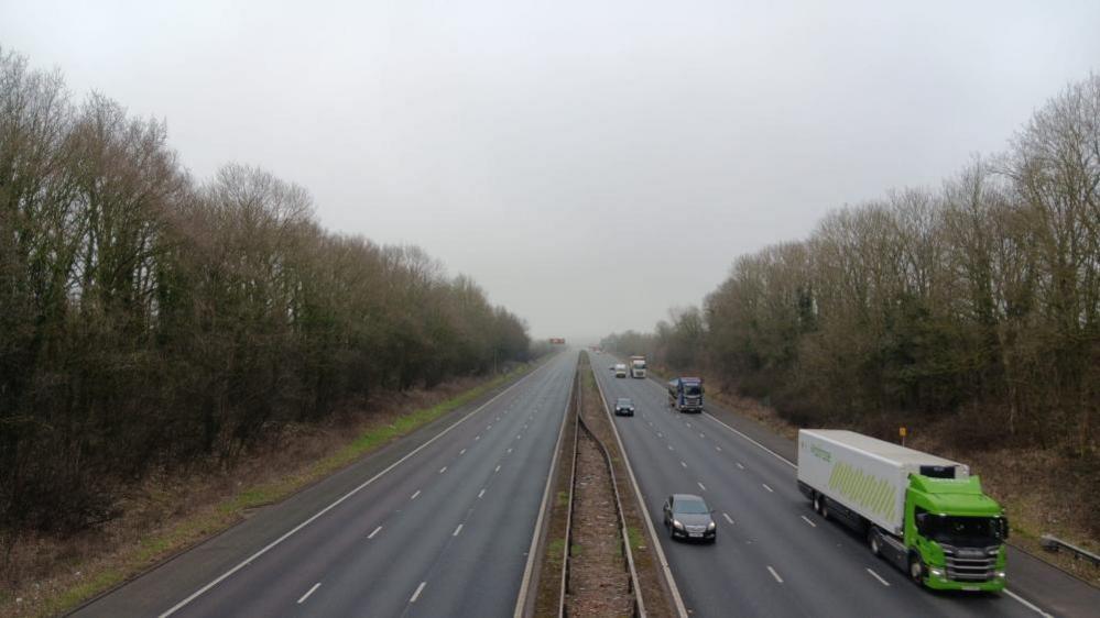 The M6 motorway with the left-hand side of the carriageway closed while the right, northbound carriage continues to have traffic. The sky is grey and there are trees on both sides of the road.  