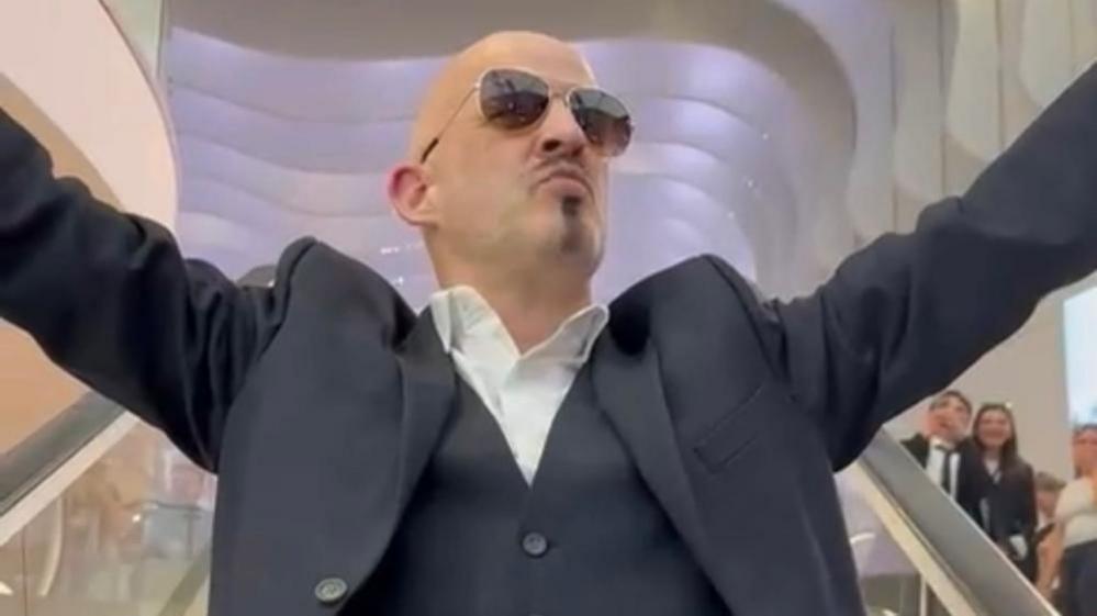 Phil Mansford dressed in sunglasses, with a white shirt, blue waistcoat, jacket, with this arms out, his lips clenched, with a bald head and dark goatee. He is on an O2 escalator, people are to the right.