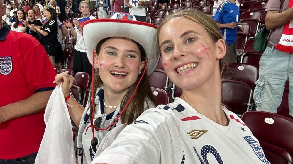 Maisie Fairweather Prescott and her friend Flo wearing white England shirts with blue insignia and three lions badges in a stadium surrounded by football fans. Natalie wears a white England cowboy style hat.