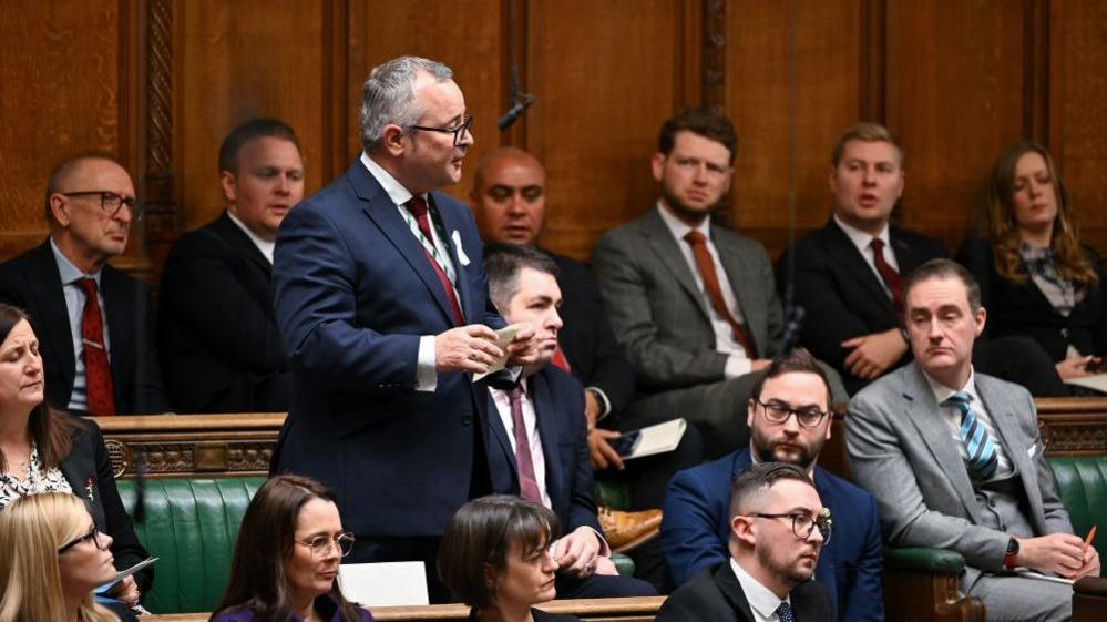 Lee Barron in a blue suit and red tie stood up in the House of Commons with other MPs sat around on the green benches