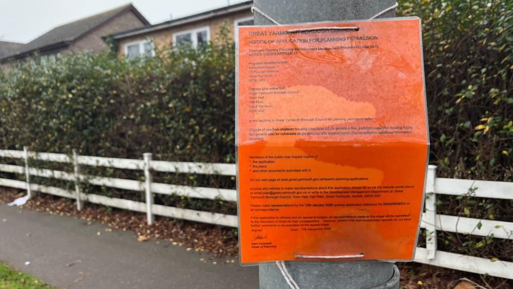 A planning notice, on orange paper is tied to a lamp post outside Abbeyfield Care Home. There is a white post and rail fence near the pavement and a tall hedge behind that, with the building visible behind that.