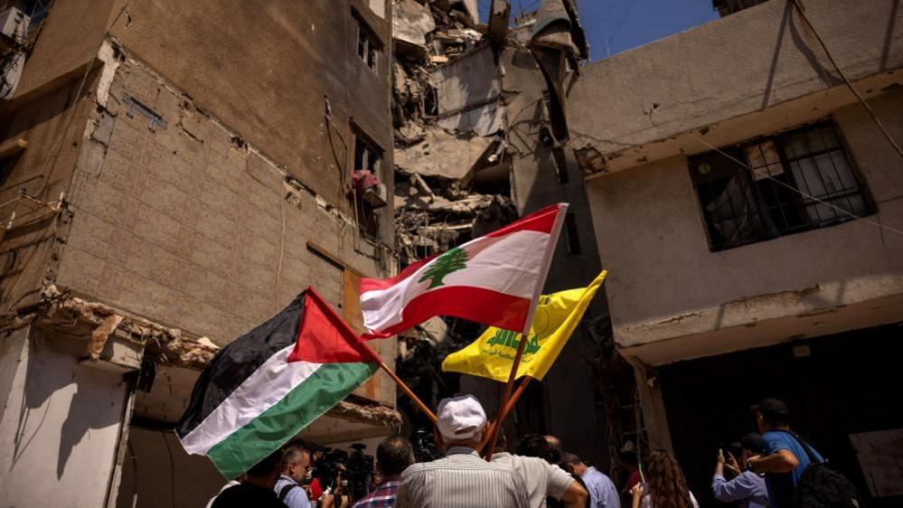 A man holds Palestinian, Lebanese and Hezbollah flags next to a damaged site where top Hezbollah commander Fuad Shukr was killed