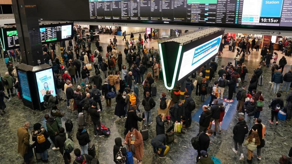 Passengers standing on a railway concourse in front of large departure screens