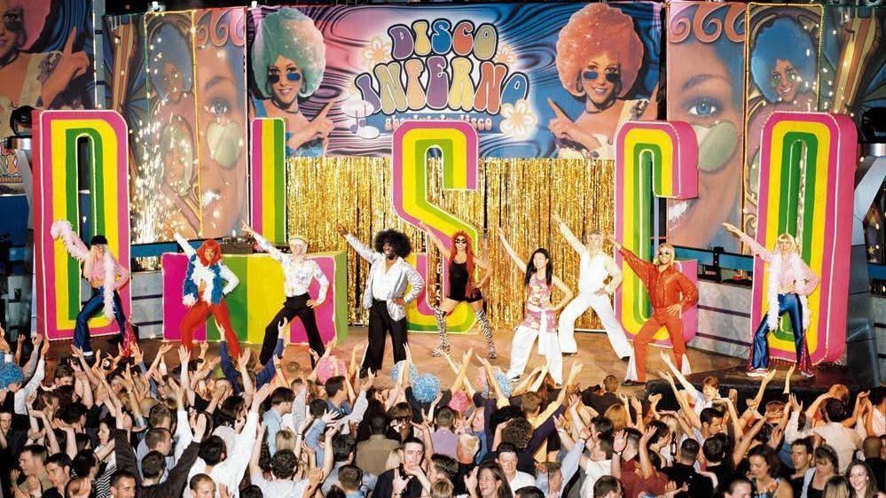 A brightly coloured stage with multicoloured letters reading "Disco" and a sign behind reading "Disco inferno" with images of women wearing brightly coloured wigs. Dancers are on the stage wearing bright 70s-style outfits and the crowd below are dancing.